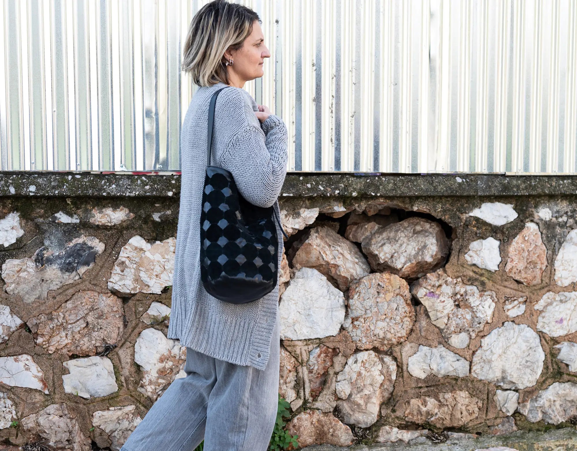 Woman holding a black patterned bag against a stone wall and metal fence background