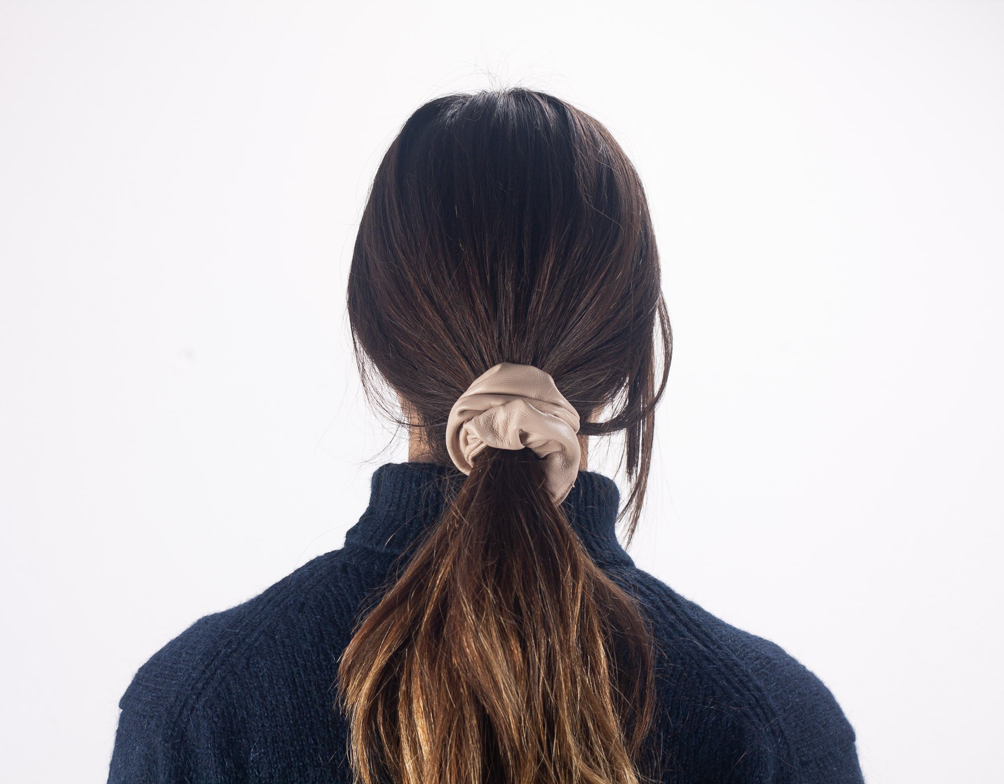 Person with long dark hair tied in a ponytail with a beige scrunchie, wearing a dark blue sweater on a white background.