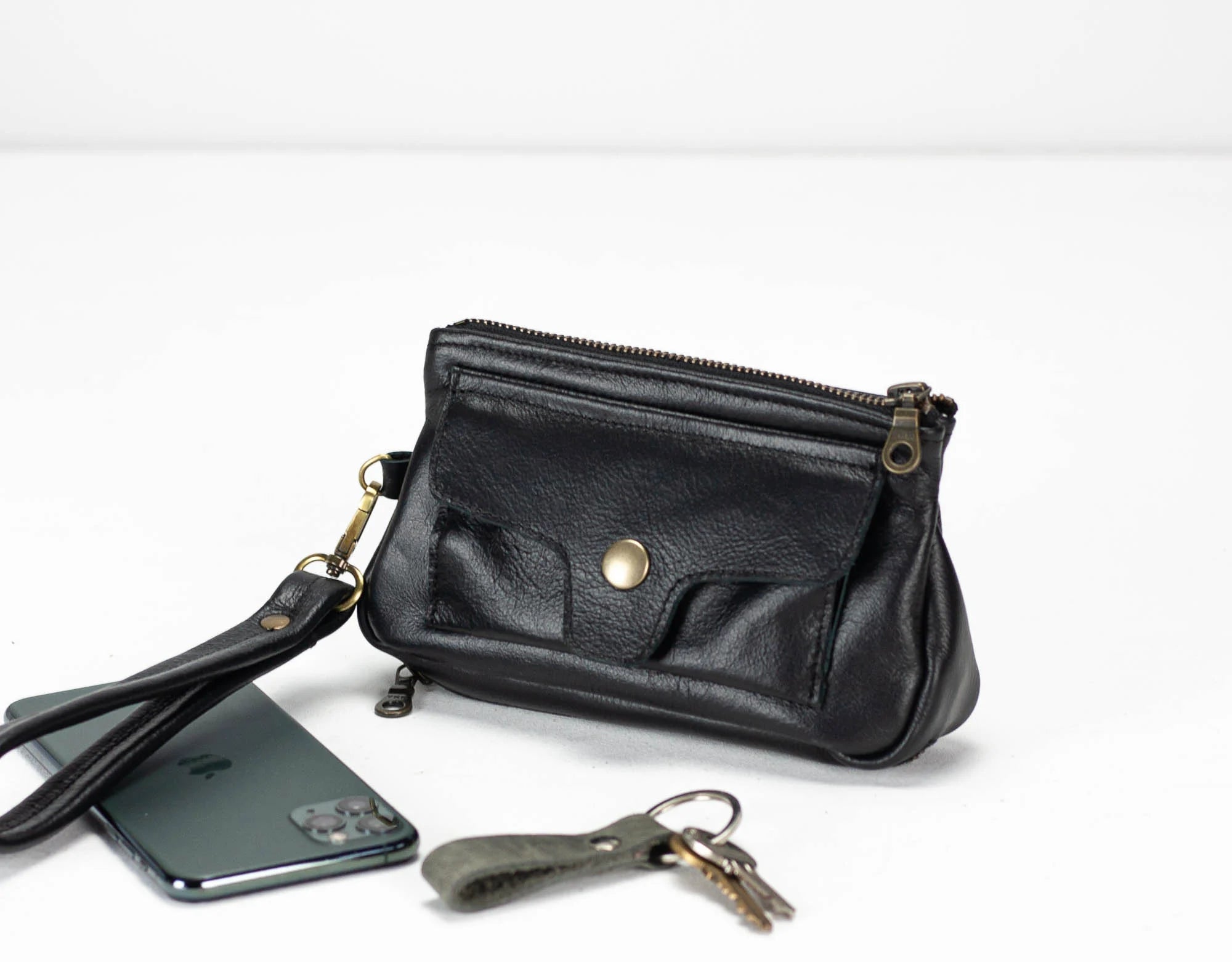 Black leather clutch with wrist strap, phone, and keys on a white background