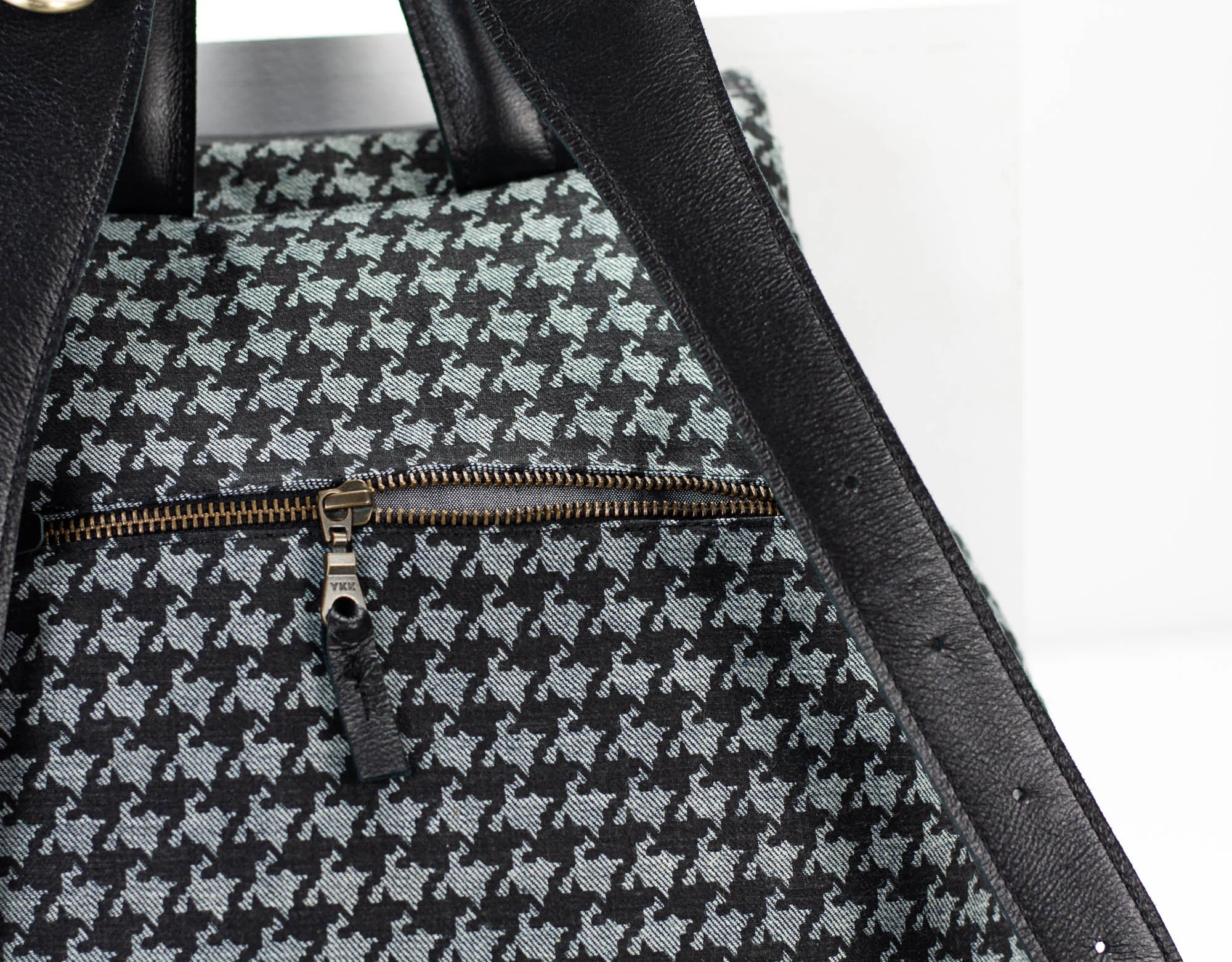 Close-up of a black and gray houndstooth patterned bag with a gold zipper on a white background.