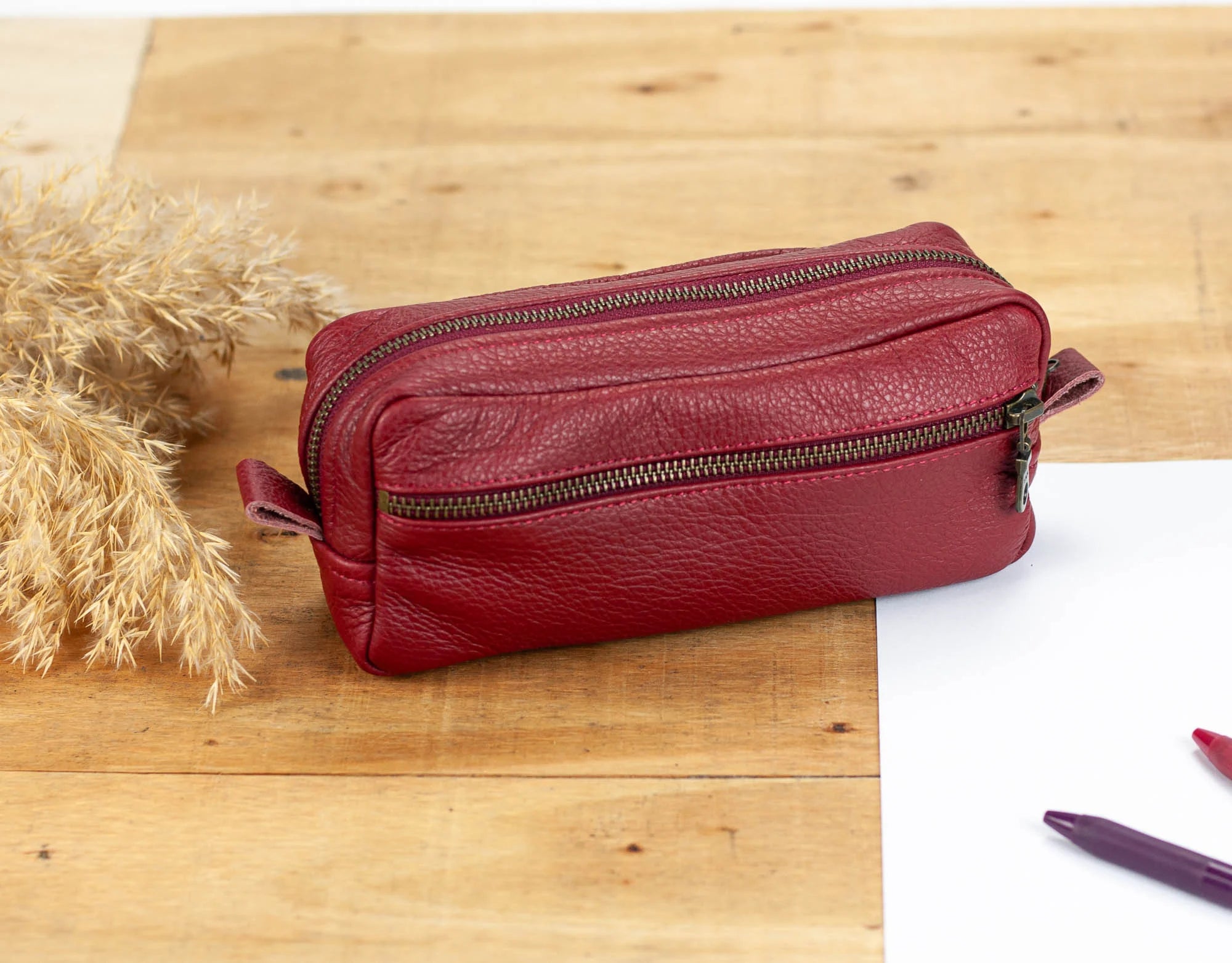 Red leather toiletry bag on a wooden surface with a white paper and pen.