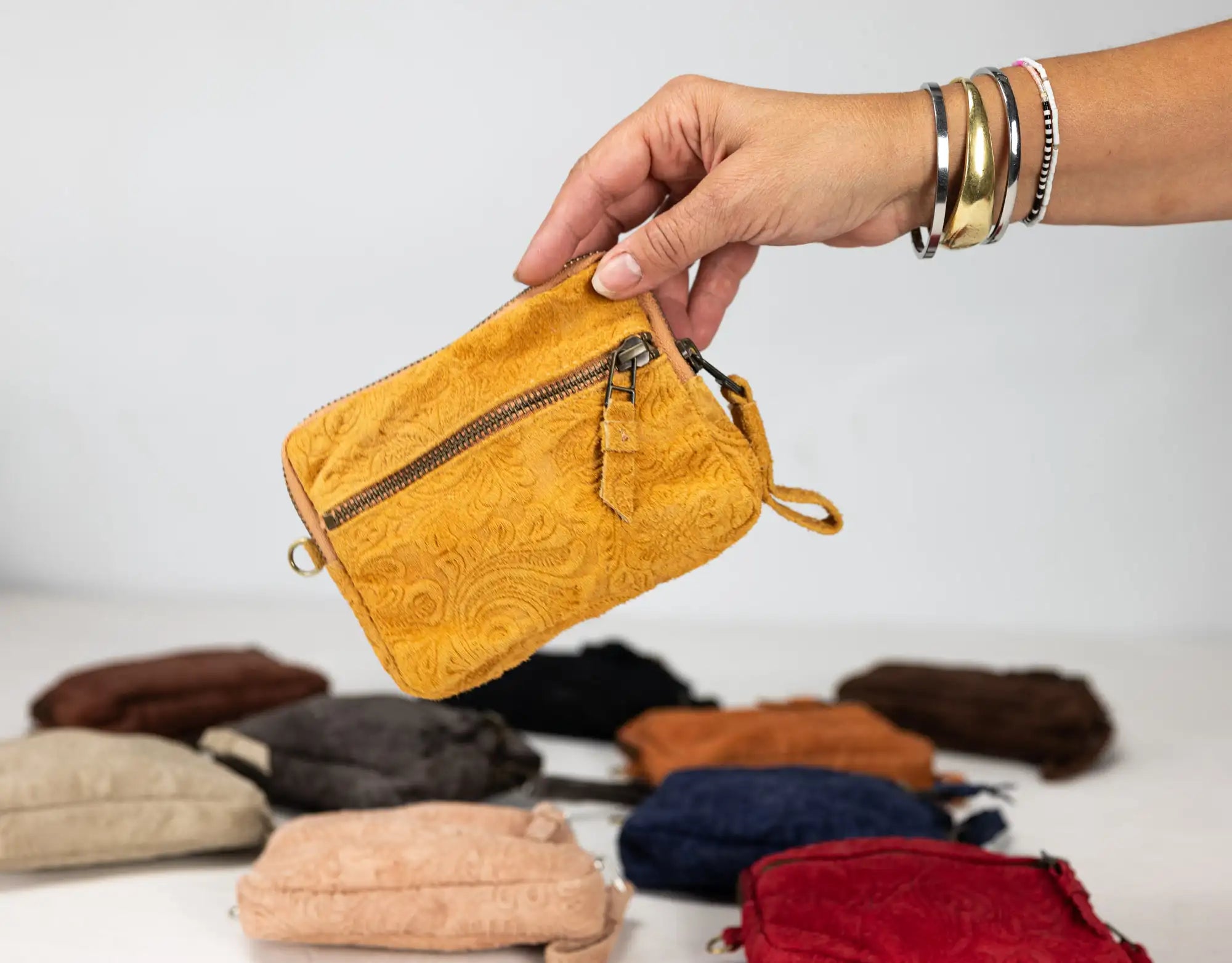 Hand holding a mustard yellow textured pouch with other pouches in the background on a light gray surface.