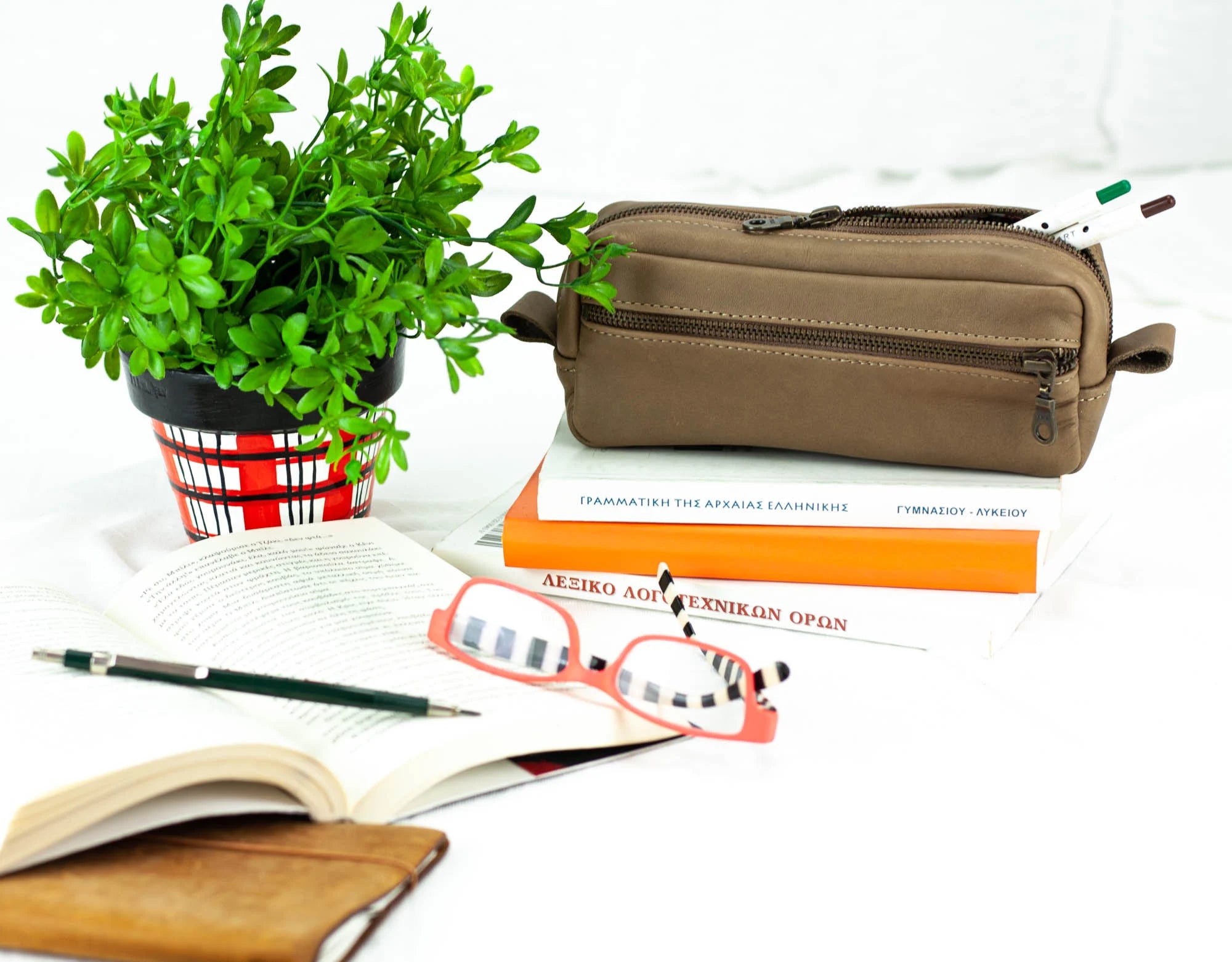 Brown toiletry bag on books with a plant, glasses, and pen on a white background
