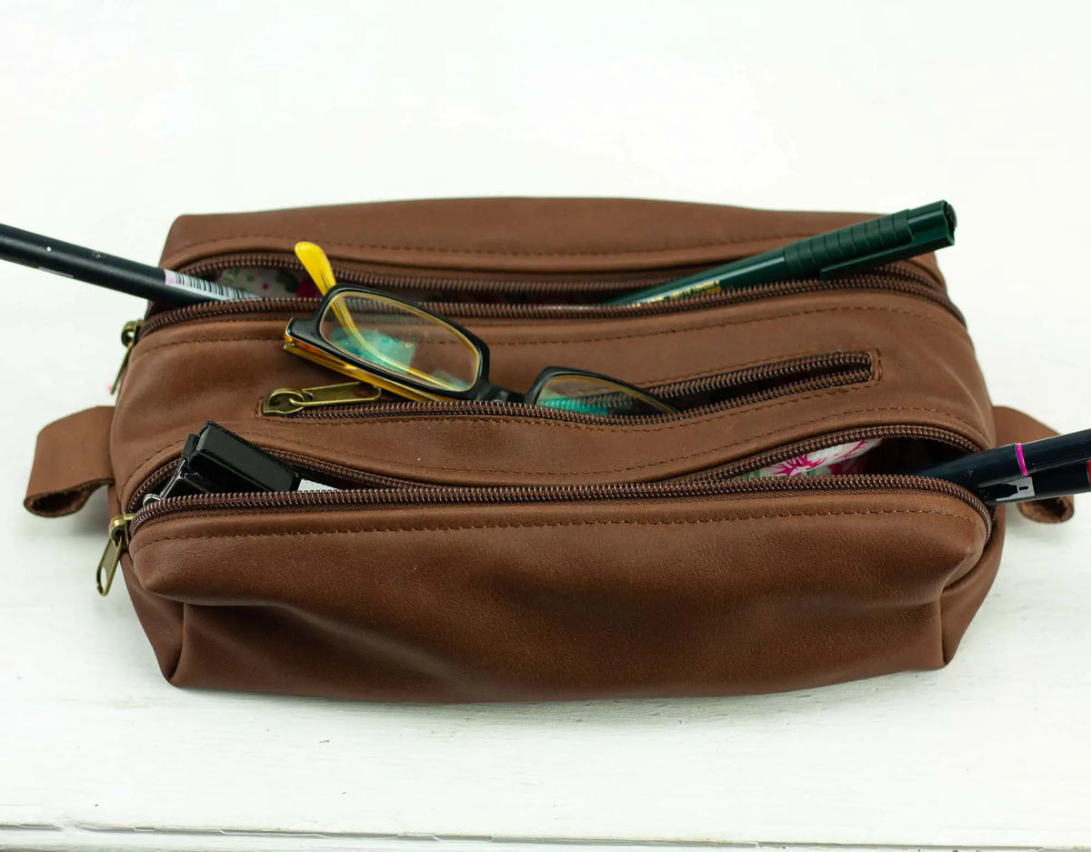 Brown leather dopp kit with glasses and other items on a white background#color_chocolate-brown