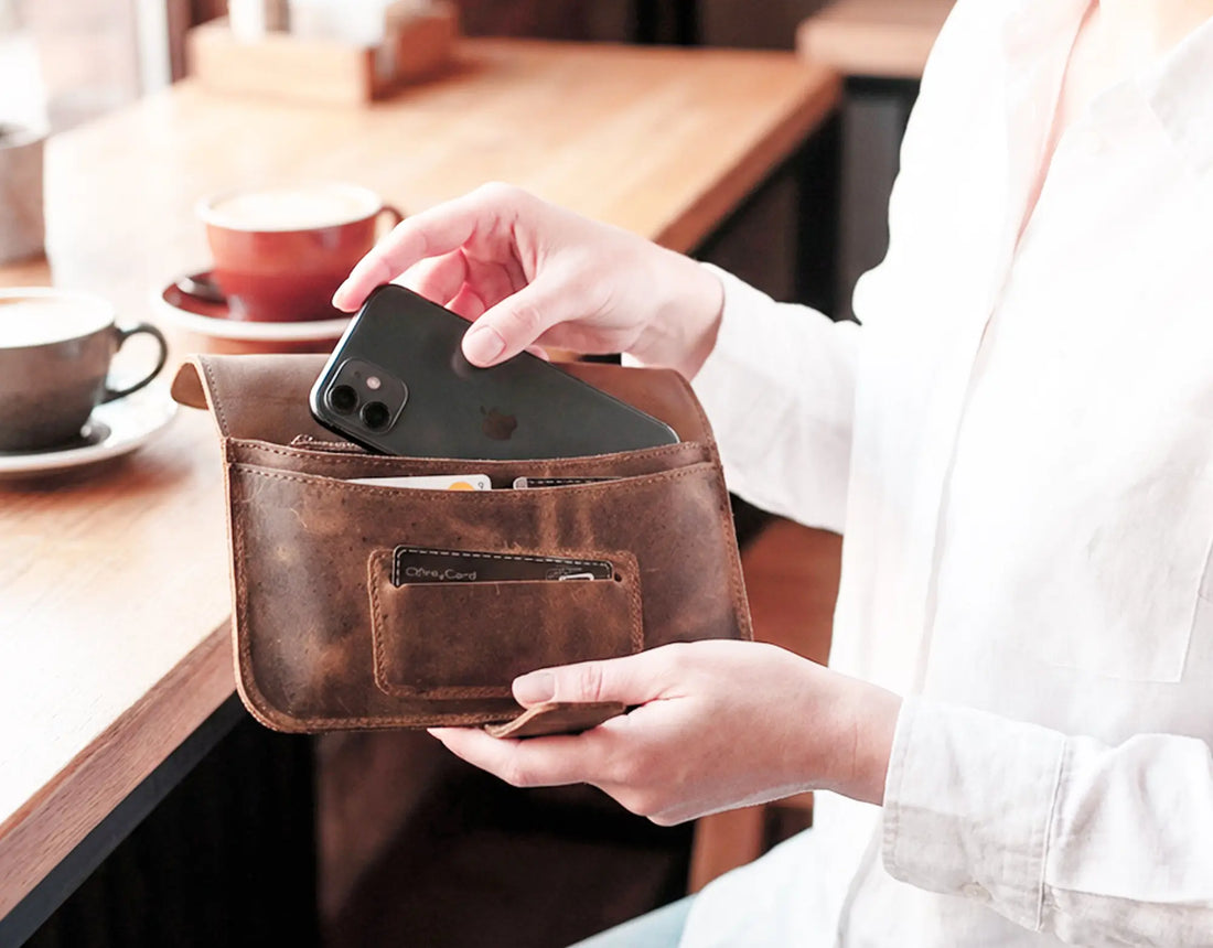 Person holding a brown leather wallet with a phone inside, sitting at a wooden table.