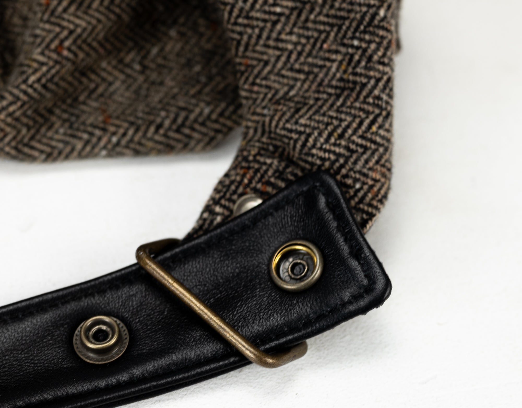 Close-up of a herringbone-patterned fabric with a black leather strap and brass buckle on a white background.