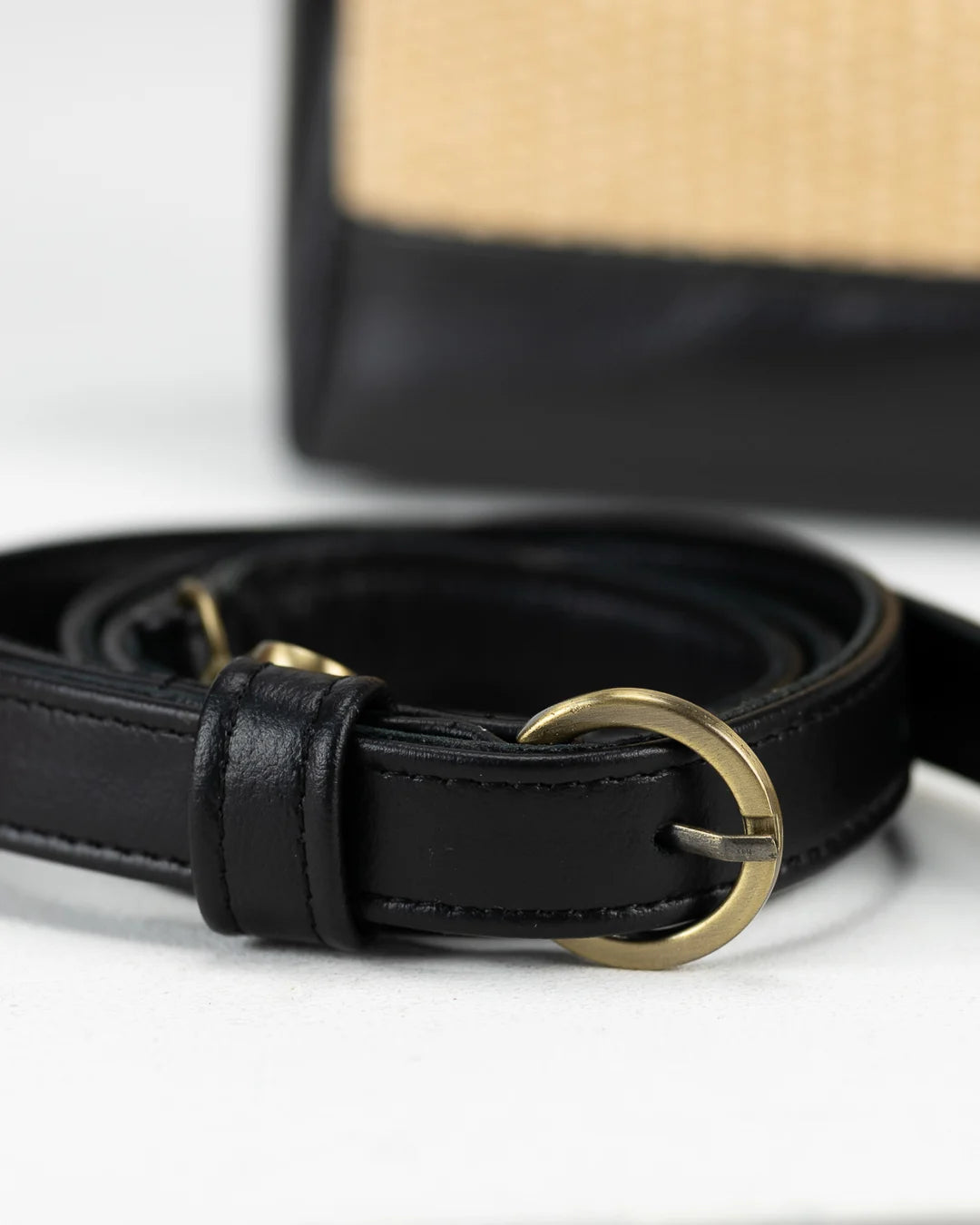 Black leather belt with a gold buckle on a white surface, with a straw hat in the background.