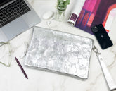 Silver metallic pouch on a marble desk with laptop, phone, and other items.
