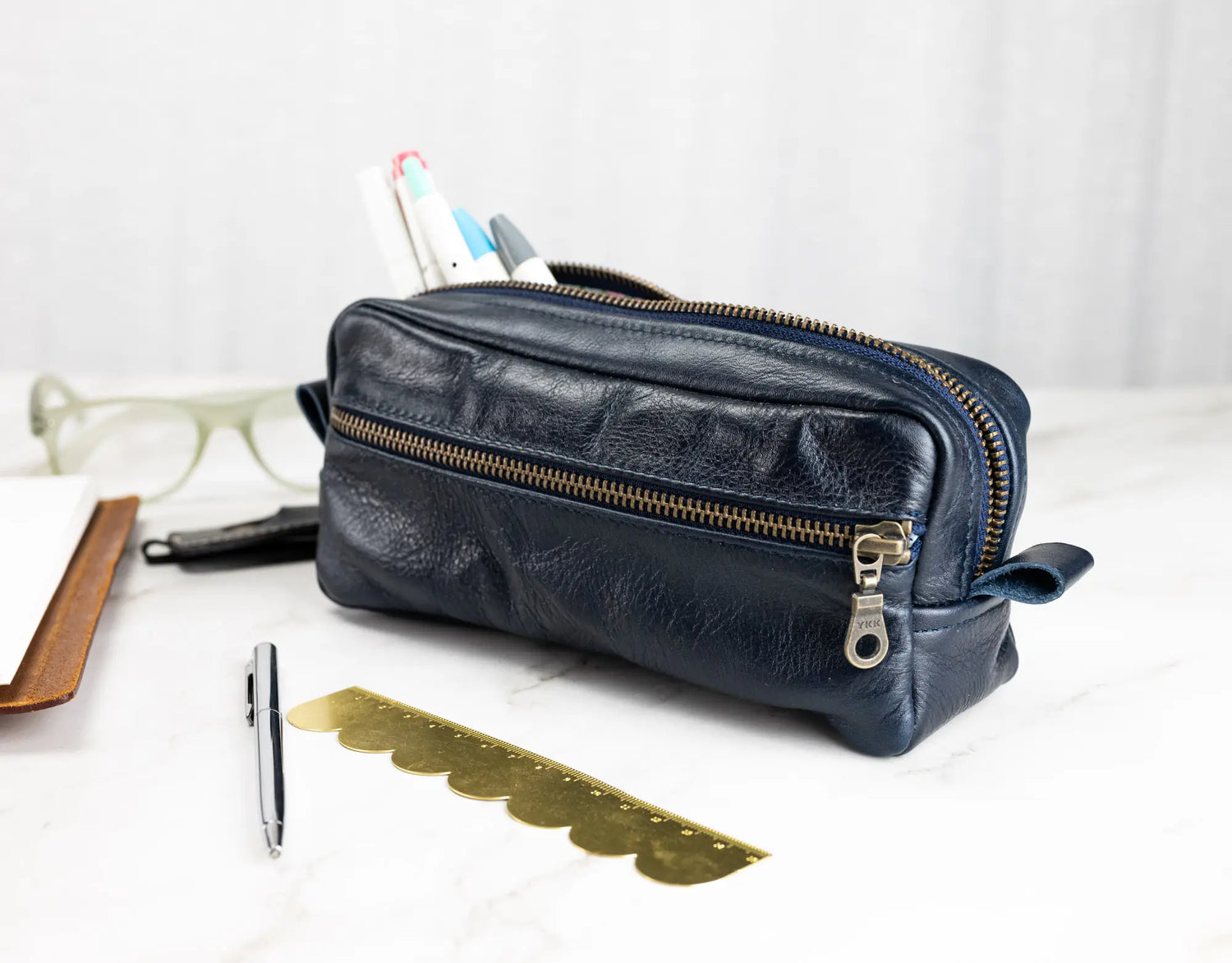 Navy blue leather dopp kit with gold zipper on a white surface with stationery items.