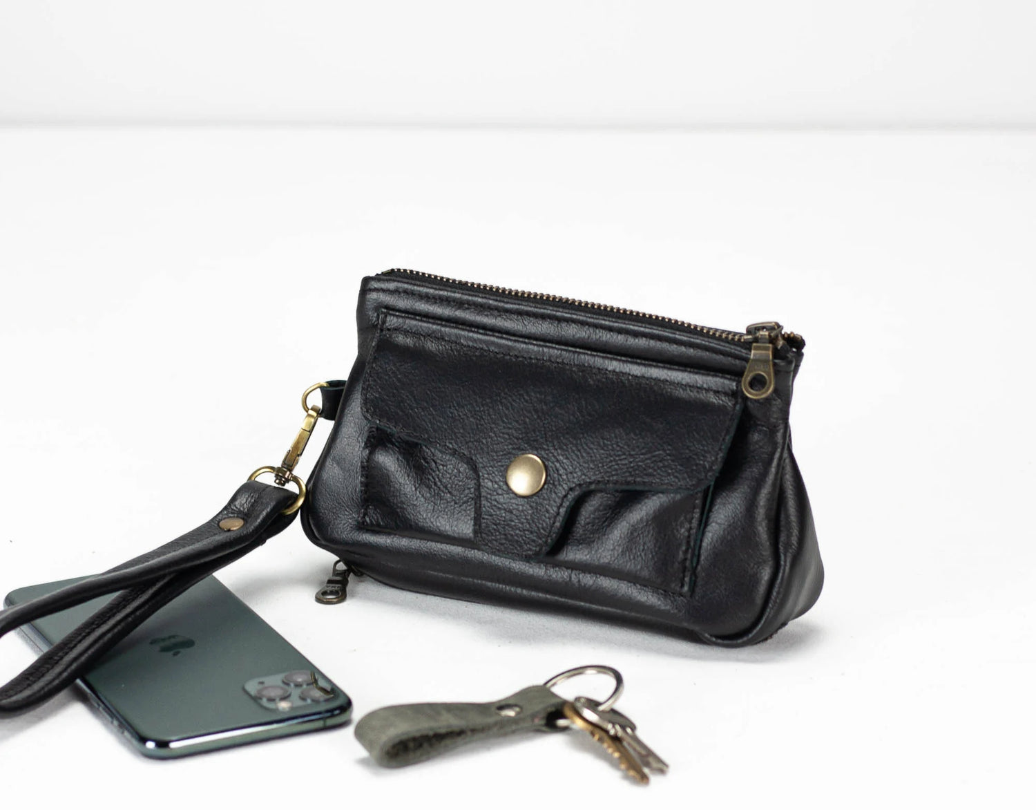 Black leather clutch with wrist strap, phone, and keys on a white background