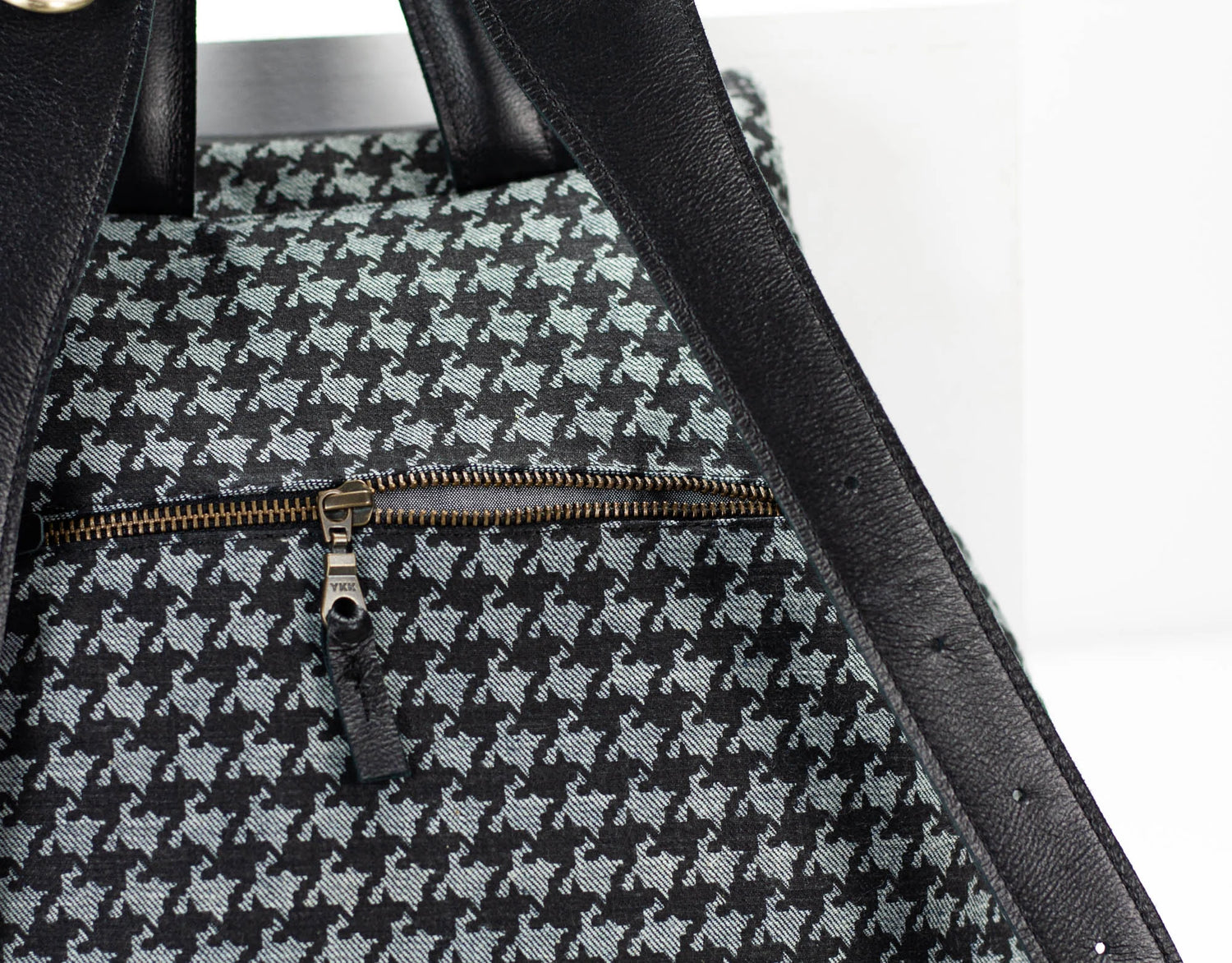Close-up of a black and gray houndstooth patterned bag with a gold zipper on a white background.