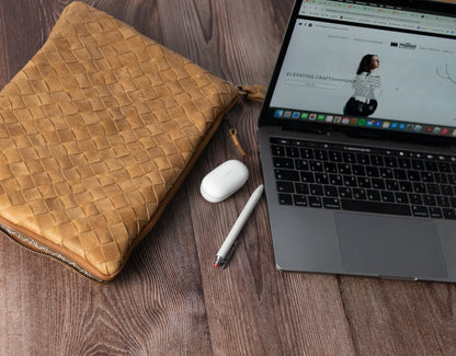 Laptop on a wooden surface with a woven brown pouch, white mouse, and pen.