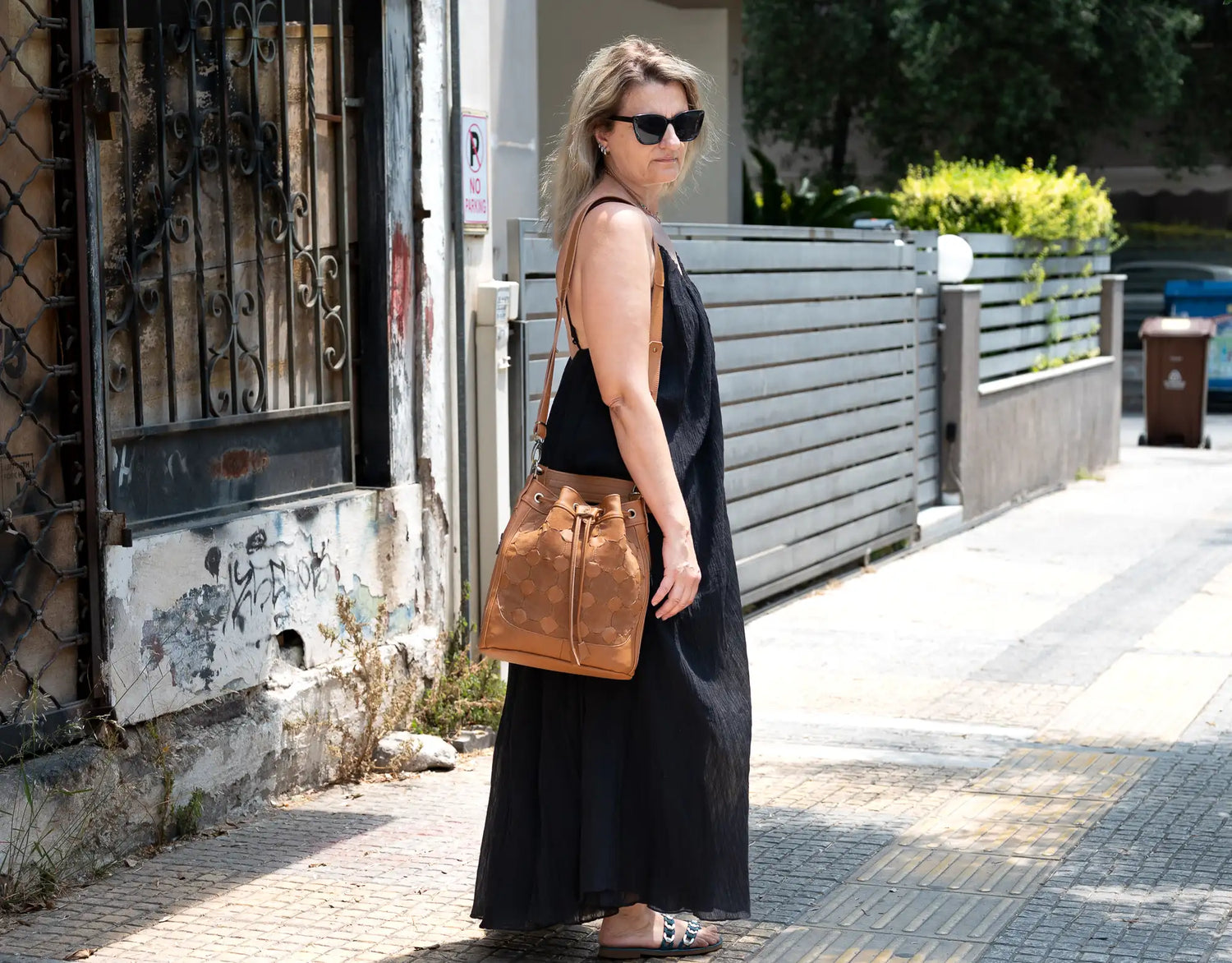 Woman in a black dress with sunglasses and a brown bag walking on a sidewalk.
