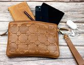 Brown leather clutch with geometric pattern on a wooden surface with phone, pen, and earbuds.
