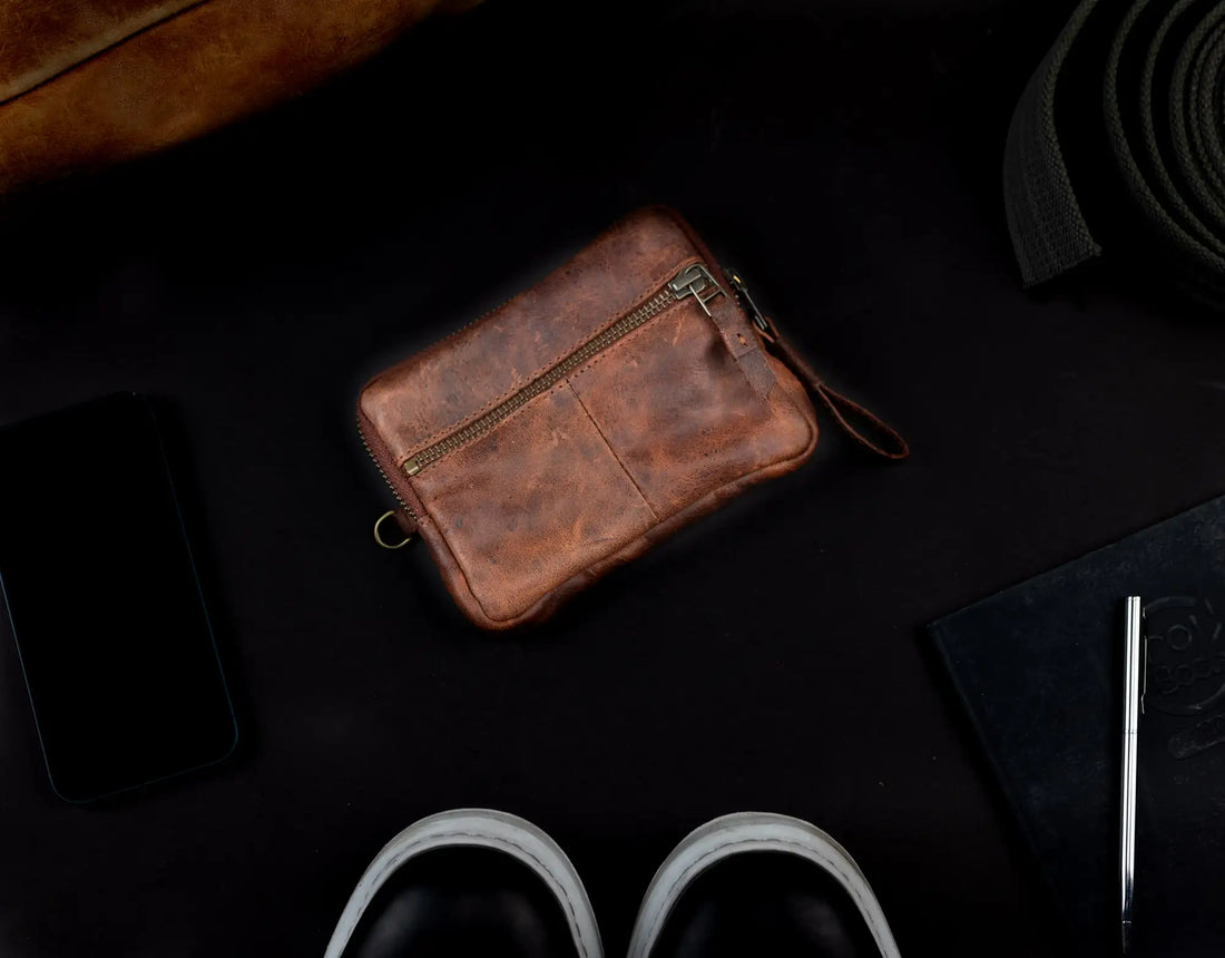 Brown leather pouch on a dark surface with a phone, slippers, and other items.