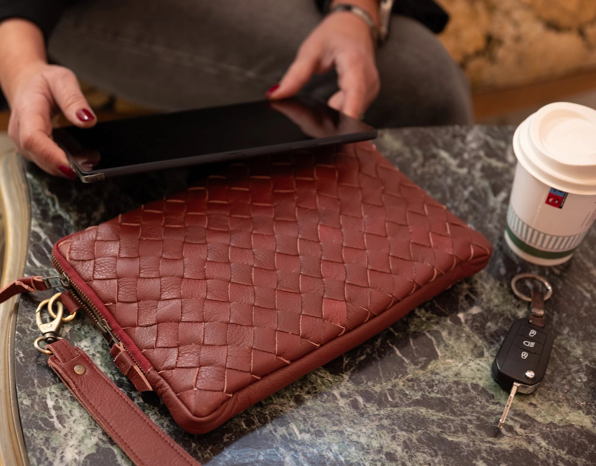 Person holding a black laptop over a woven brown leather clutch on a marble surface with a coffee cup and keys.