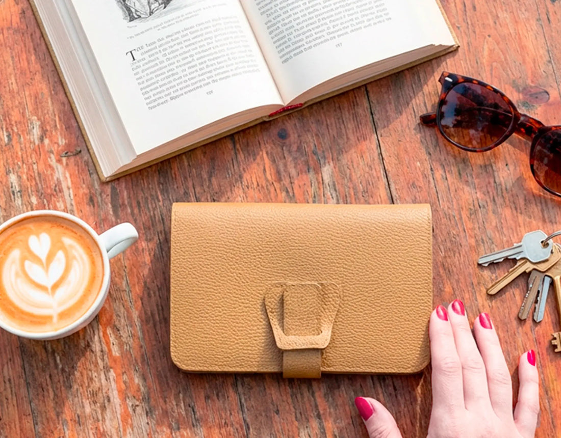 Brown leather wallet on a wooden table with a book, sunglasses, and coffee.