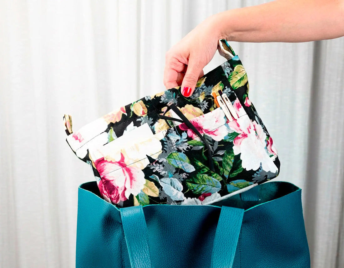 Hand holding a floral clutch over a blue bag with a white curtain background