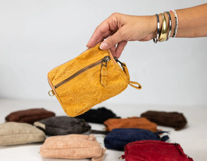 Hand holding a mustard yellow textured pouch with other pouches in the background on a light gray surface.