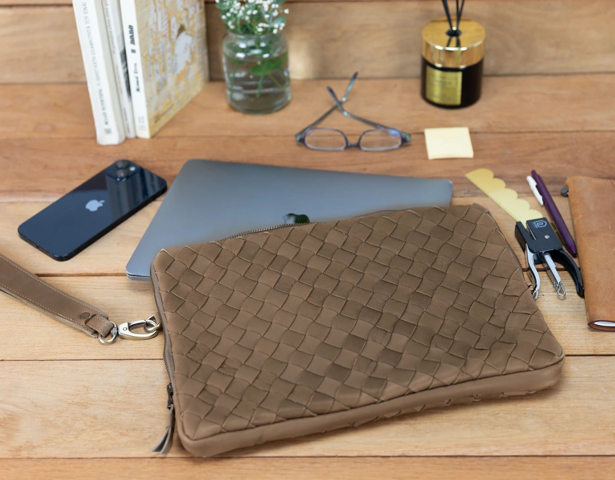 Brown woven clutch bag on a wooden desk with phone, laptop, and other items.