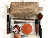 Makeup items including a Clarins product, lip gloss, and brushes on a marble surface with a woven beige pouch.