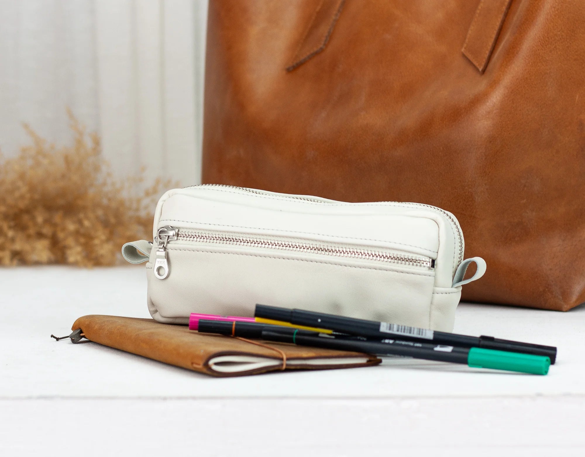 White pencil case with pens and a brown leather bag on a light surface
