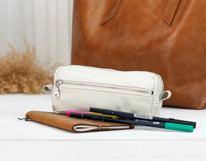 White pencil case with pens and a brown leather bag on a light surface