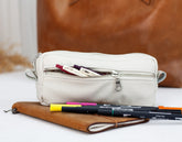White pencil case with pens on a white surface, with a brown leather bag in the background.