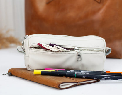 White pencil case with pens on a white surface, with a brown leather bag in the background.