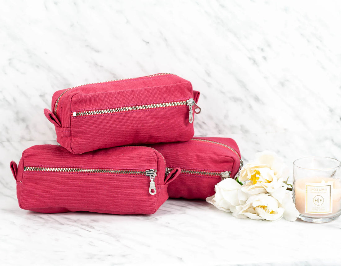 Three pink toiletry bags with zippers on a white textured surface with a candle and flowers.