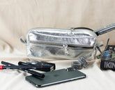 Silver fanny pack with makeup items and a phone on a beige surface