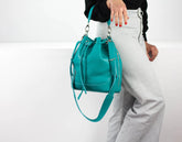 Teal handbag held by a person wearing white pants on a light gray background