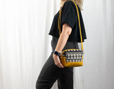 Person holding a mustard yellow crossbody bag with a patterned pocket against a white curtain background
