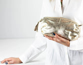 Gold clutch bag held by a person wearing a white outfit on a light gray background
